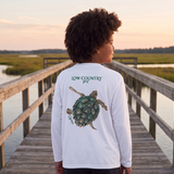 Person standing on a wooden dock by a body of water, wearing a white long-sleeve shirt with a turtle design.